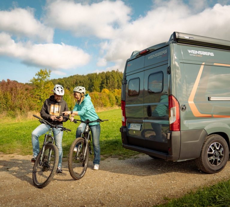 Campingplatz Marketing Zwei Personen mit Fahrrädern stehen neben einem Wohnmobil im Freien.