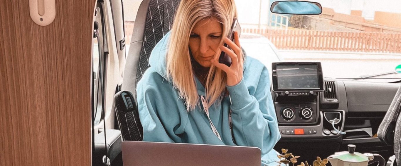 Frau mit blonden Haaren sitzt im Fahrzeug und spricht am Telefon, vor ihr ein Laptop.